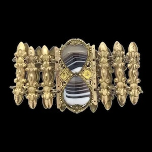 Könnte beinhalten: Ein goldenes Armband mit einem dekorativen Design und einem großen, ovalen Achat-Stein in der Mitte. Der Stein hat ein gestreiftes Muster in Braun-, Weiß- und Grautönen.