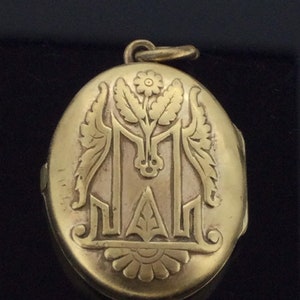 K&ouml;nnte beinhalten: Ein ovales goldenes Medaillon mit einem gravierten Blumendesign und dem Buchstaben "M".