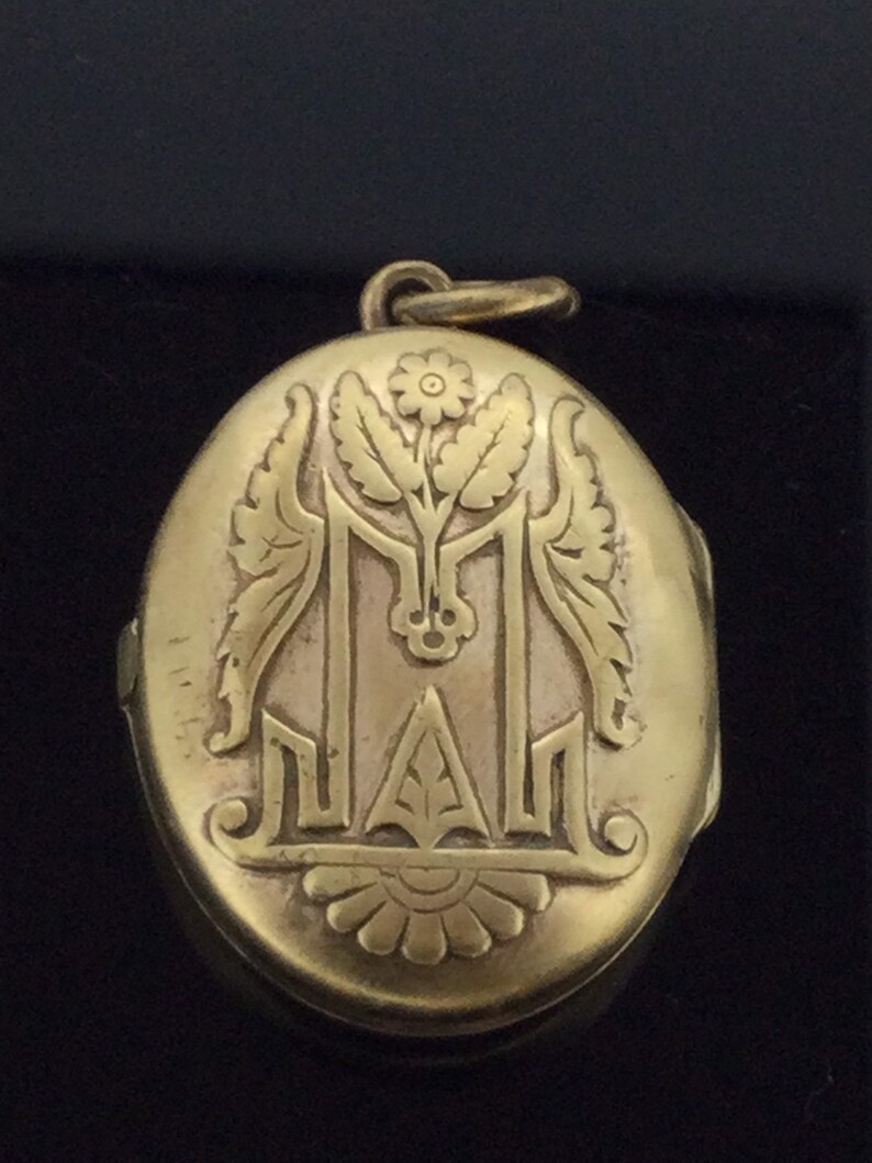 K&ouml;nnte beinhalten: Ein ovales goldenes Medaillon mit einem gravierten Blumendesign und dem Buchstaben "M".