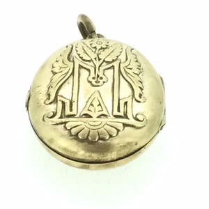 K&ouml;nnte beinhalten: Ein goldenes Medaillon mit einem gravierten Blumendesign und dem Buchstaben "M" in der Mitte.