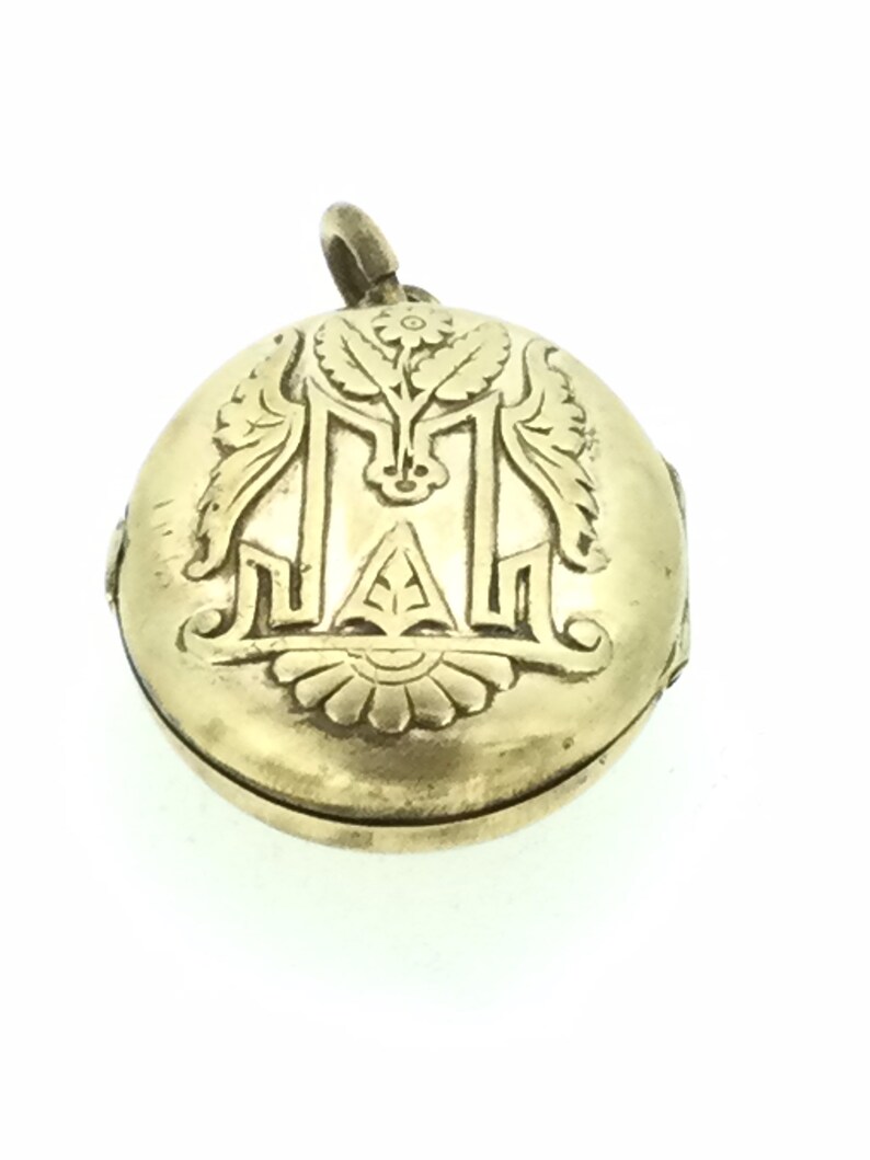 K&ouml;nnte beinhalten: Ein goldenes Medaillon mit einem gravierten Blumendesign und dem Buchstaben "M" in der Mitte.
