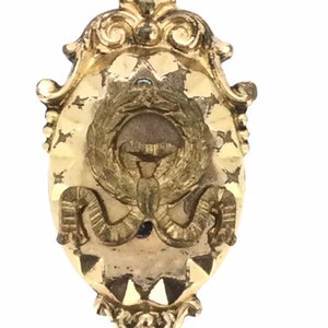 May include: A gold-toned metal brooch with an ornate design. The center features a stylized image of a bird with wings spread wide. The brooch is oval shaped and has a decorative border.