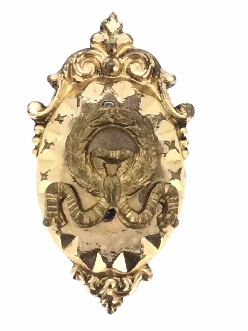 May include: A gold-toned metal brooch with an ornate design. The center features a stylized image of a bird with wings spread wide. The brooch is oval shaped and has a decorative border.