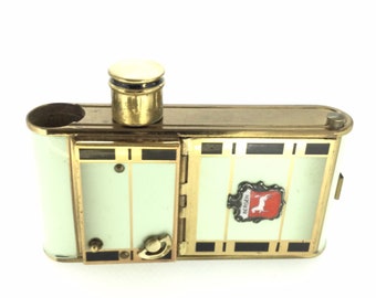 May include: A gold and white metal cigarette case with a hinged lid and a small, round, gold-colored top. The case has a black and gold geometric design and a small, white enamel emblem with the word "BERGEN" and a red and white image of a horse.