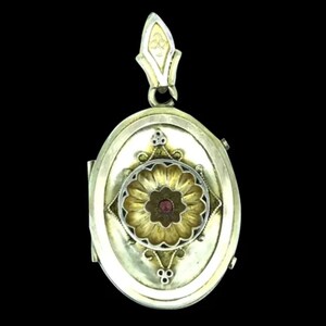 Pode incluir: Um medalhão oval de prata com um design floral dourado e uma pedra preciosa vermelha no centro. O medalhão tem uma tampa com dobradiças e um laço na parte superior para prender uma corrente.