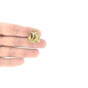 May include: A gold-colored pendant in the shape of a ship with intricate details. The pendant is small and delicate, and it is held in a person's hand.