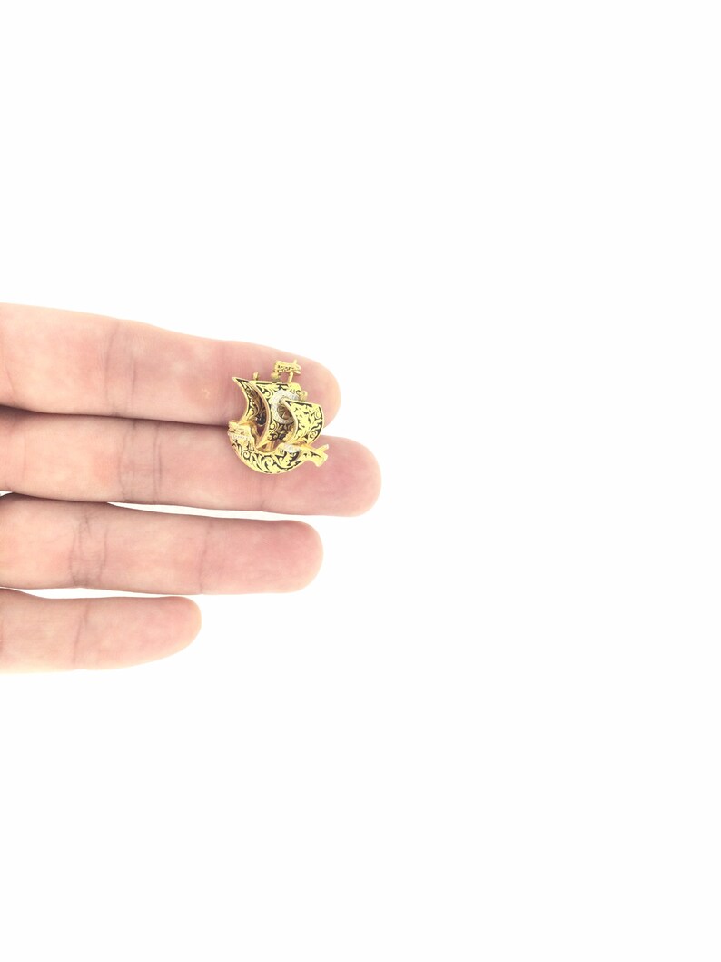 May include: A gold-colored pendant in the shape of a ship with intricate details. The pendant is small and delicate, and it is held in a person's hand.