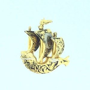 May include: A gold-colored ship pendant with black enamel detailing. The ship has a sail and a flag. The pendant is ornate and detailed.