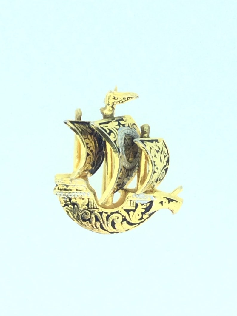 May include: A gold-colored ship pendant with black enamel detailing. The ship has a sail and a flag. The pendant is ornate and detailed.