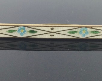 Puede incluir: Un broche de barra de esmalte blanco con un diseño geométrico con flores azules y verdes. El broche es plateado y tiene una forma rectangular.