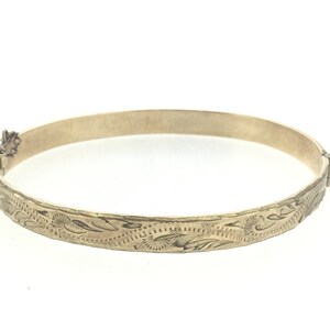 K&ouml;nnte beinhalten: Ein goldenes Armreif-Armband mit einem floralen, gravierten Design.
