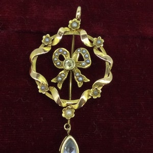 May include: A gold pendant with a pearl and diamond bow design, hanging from a gold chain with a teardrop-shaped blue gemstone.