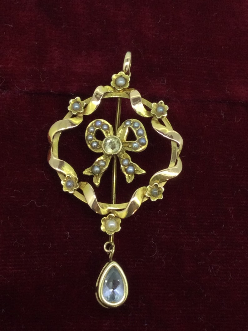 May include: A gold pendant with a pearl and diamond bow design, hanging from a gold chain with a teardrop-shaped blue gemstone.