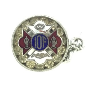 May include: Silver and blue enamel pin with the letters "IOF" in the center, surrounded by a red and white cross with a stylized eye at the top. The pin has a chain attached to the back.