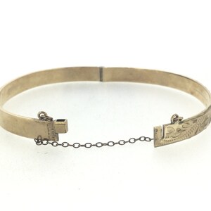 K&ouml;nnte beinhalten: Ein goldenes Armreif-Armband mit einer zarten Kette und einem Verschluss. Das Armband ist mit einem Blumenmuster graviert.