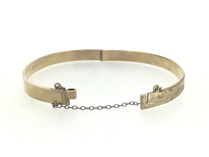K&ouml;nnte beinhalten: Ein goldenes Armreif-Armband mit einer zarten Kette und einem Verschluss. Das Armband ist mit einem Blumenmuster graviert.