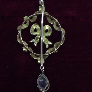 May include: A gold pendant with a teardrop-shaped stone hanging from a circular frame with a bow design. The frame is decorated with small stones.