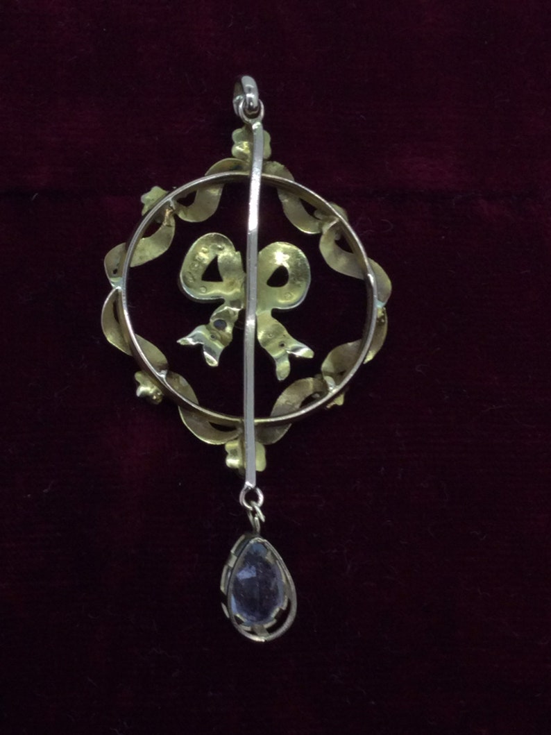 May include: A gold pendant with a teardrop-shaped stone hanging from a circular frame with a bow design. The frame is decorated with small stones.