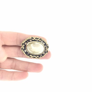 May include: A gold-colored oval brooch with a black enamel background and a white cameo. The brooch is decorated with gold filigree and the words "Memento Mori" are inscribed around the edge.