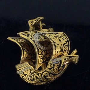 May include: A gold-colored metal ship pendant with black enamel detailing. The ship has intricate floral designs and a cross on the sail.