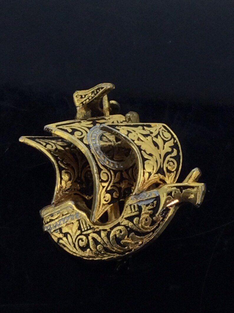 May include: A gold-colored metal ship pendant with black enamel detailing. The ship has intricate floral designs and a cross on the sail.