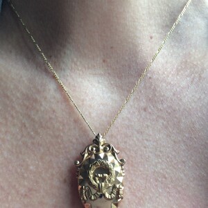 May include: A gold chain necklace with a detailed gold pendant featuring a wreath and a stylized figure. The pendant is oval shaped and has a textured surface.