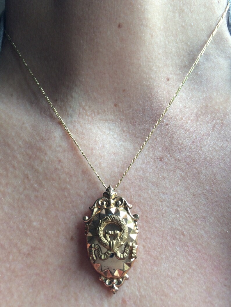 May include: A gold chain necklace with a detailed gold pendant featuring a wreath and a stylized figure. The pendant is oval shaped and has a textured surface.