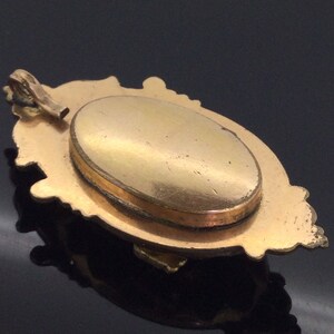 May include: A gold-toned locket with an ornate design and a hinged oval opening. The locket has a smooth, polished surface and a delicate chain attached to the top.