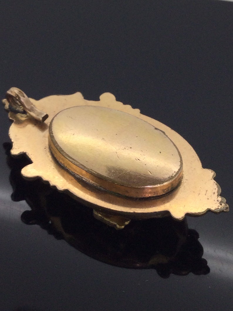 May include: A gold-toned locket with an ornate design and a hinged oval opening. The locket has a smooth, polished surface and a delicate chain attached to the top.