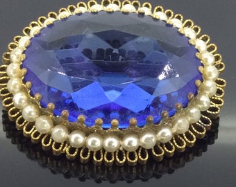 May include: A vintage brooch with a large, faceted, blue glass stone surrounded by a gold-tone setting and a row of faux pearls.