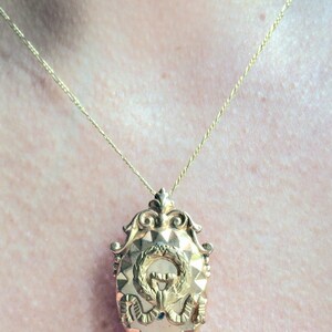 May include: A gold chain necklace with a gold pendant featuring a wreath and a monogram. The pendant is oval shaped and has a textured surface.