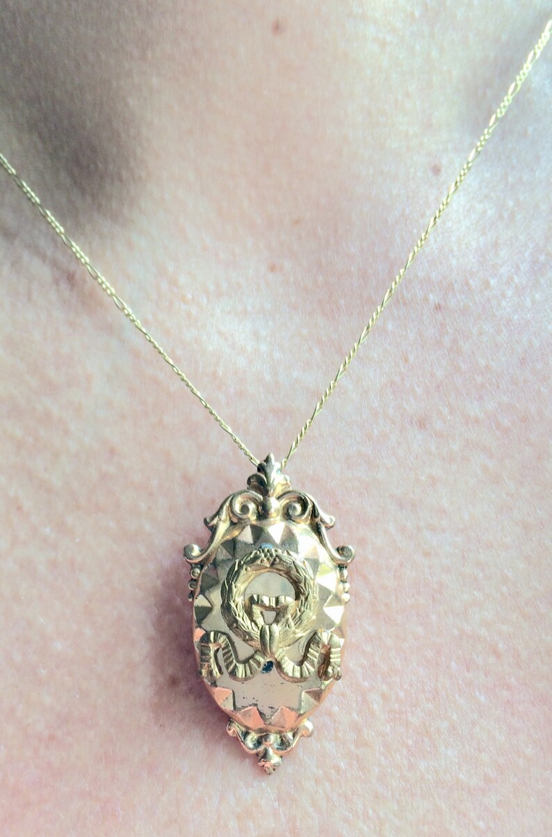 May include: A gold chain necklace with a gold pendant featuring a wreath and a monogram. The pendant is oval shaped and has a textured surface.
