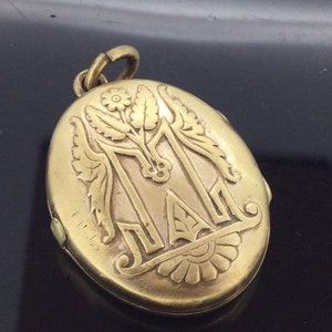 K&ouml;nnte beinhalten: Goldenes ovales Medaillon mit einem gravierten Blumendesign und dem Buchstaben "M" in der Mitte.