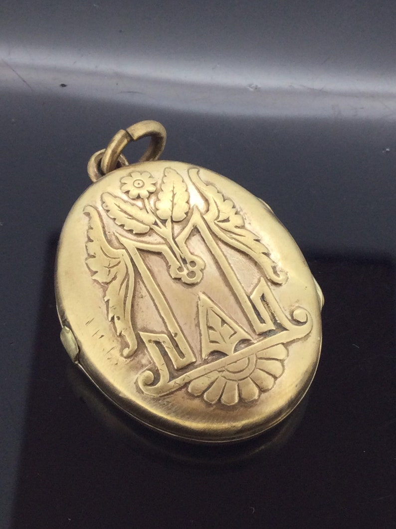 K&ouml;nnte beinhalten: Goldenes ovales Medaillon mit einem gravierten Blumendesign und dem Buchstaben "M" in der Mitte.