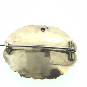 May include: The back of a gold-toned oval brooch with a pin and clasp. The brooch has a textured, scalloped edge.