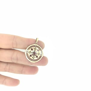 以下が含まれることがあります： 中央に赤い宝石、周囲に小さな白い宝石が散りばめられた円形のデザインの金色のペンダント