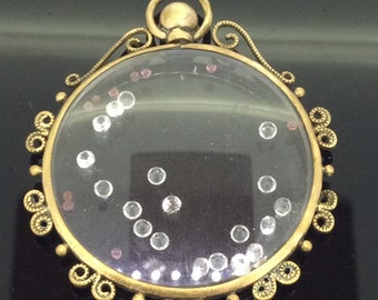 May include: A gold locket with a clear glass front and a decorative filigree border. Inside the locket are small, clear crystals and a few pink crystals.