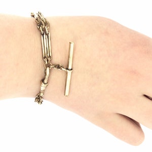Könnte beinhalten: Eine goldfarbene Taschenuhrkette mit einem T-Bar-Verschluss. Die Kette besteht aus mehreren Strängen miteinander verbundenen Metalls.