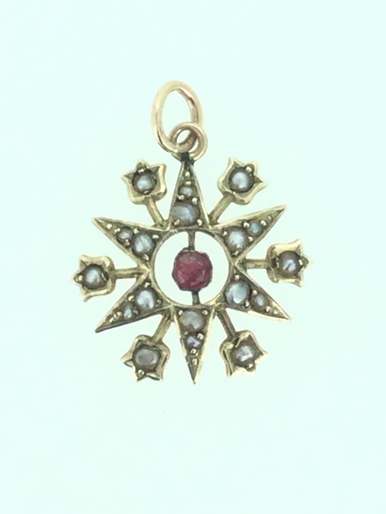 May include: A gold pendant with a red gemstone in the center, surrounded by a starburst design with small white pearls.