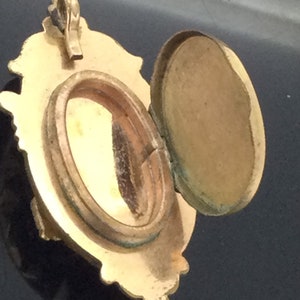May include: A gold-toned locket with an ornate design and a hinged lid. The locket is open, revealing an empty oval-shaped interior.
