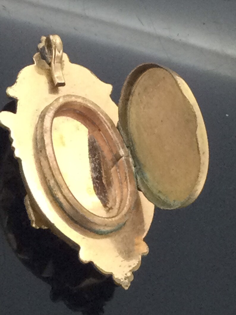 May include: A gold-toned locket with an ornate design and a hinged lid. The locket is open, revealing an empty oval-shaped interior.