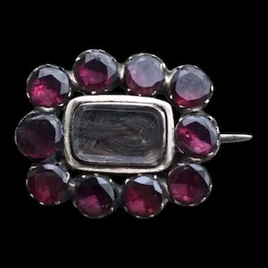 May include: A silver brooch with a rectangular glass locket containing a lock of hair surrounded by a circle of small, dark red gemstones.