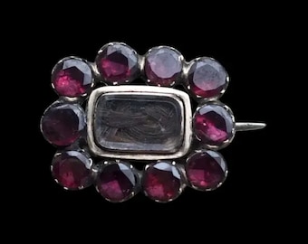 May include: A silver brooch with a rectangular glass locket containing a lock of hair surrounded by a circle of small, dark red gemstones.
