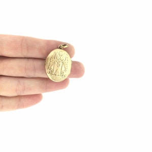 K&ouml;nnte beinhalten: Ein ovales goldenes Medaillon mit einem gravierten Design, das zwei Figuren und den Buchstaben "M" zeigt.