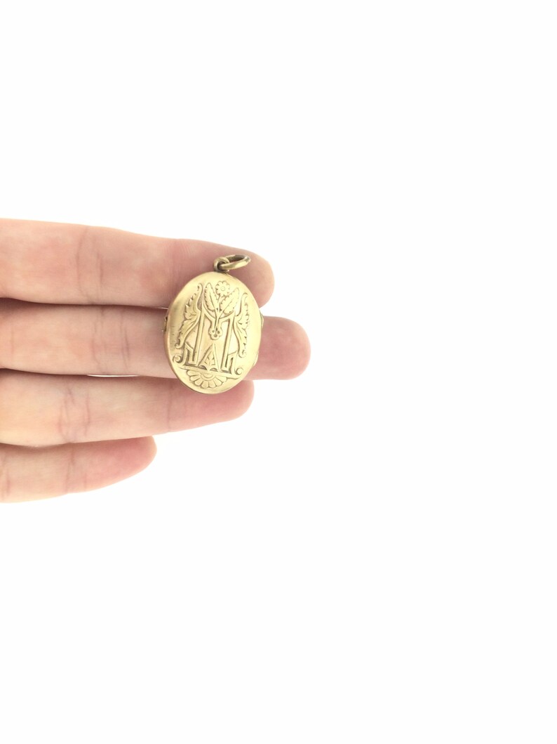 K&ouml;nnte beinhalten: Ein ovales goldenes Medaillon mit einem gravierten Design, das zwei Figuren und den Buchstaben "M" zeigt.