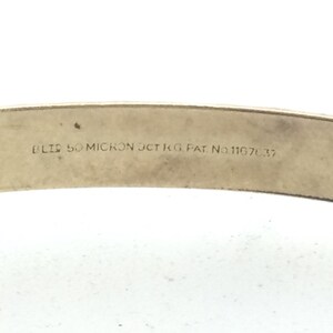 K&ouml;nnte beinhalten: Nahaufnahme eines goldfarbenen Metallarmbands mit der Inschrift "BLIS 50 MICRON JCT KG PAT. NO. 111670.37" auf der Innenseite.