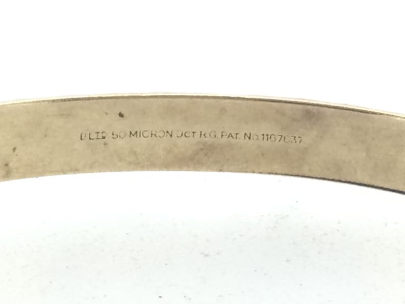 K&ouml;nnte beinhalten: Nahaufnahme eines goldfarbenen Metallarmbands mit der Inschrift "BLIS 50 MICRON JCT KG PAT. NO. 111670.37" auf der Innenseite.