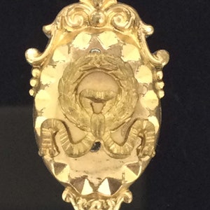 May include: A gold-toned oval-shaped brooch with a decorative design. The center of the brooch features a wreath with a stylized image of a bird inside.