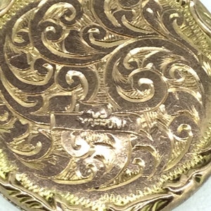May include: A gold locket with an intricate engraved design of swirling patterns. The locket has a smooth, rounded surface and a slightly raised edge.