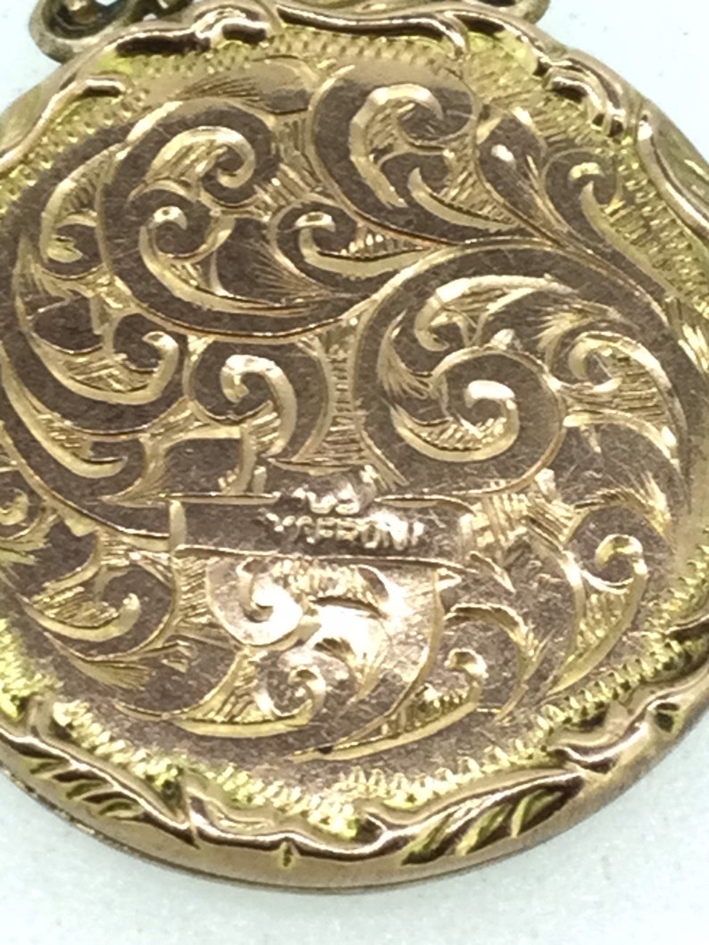 May include: A gold locket with an intricate engraved design of swirling patterns. The locket has a smooth, rounded surface and a slightly raised edge.